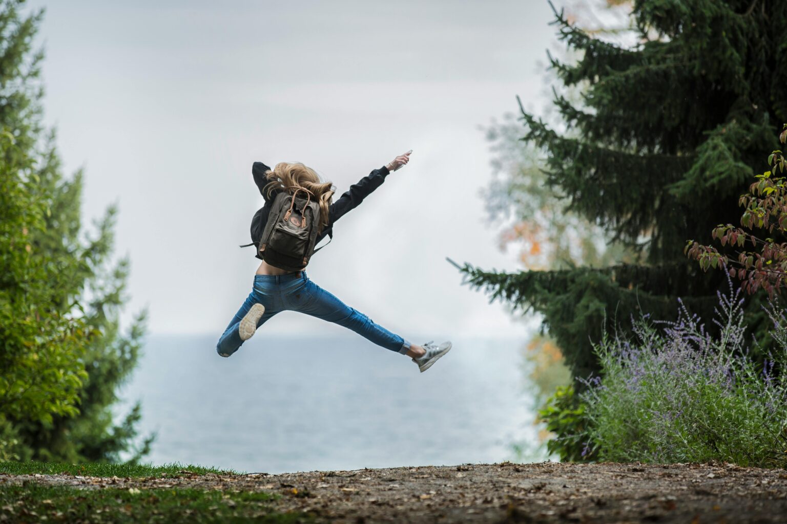 Energetic woman leaping in outdoor forest setting with lake view, showcasing freedom and joy.