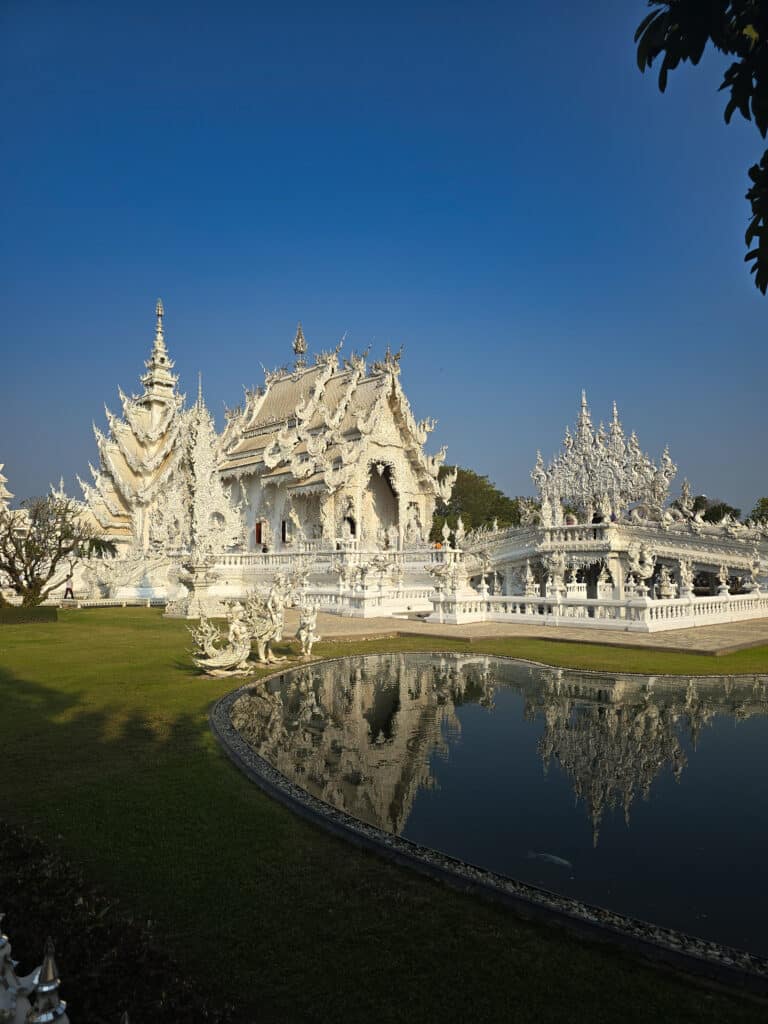 Vue du Wat Rong Khun, le célèbre Temple Blanc de Chiang Rai en Thaïlande, se reflétant dans un bassin sous un ciel bleu clair.