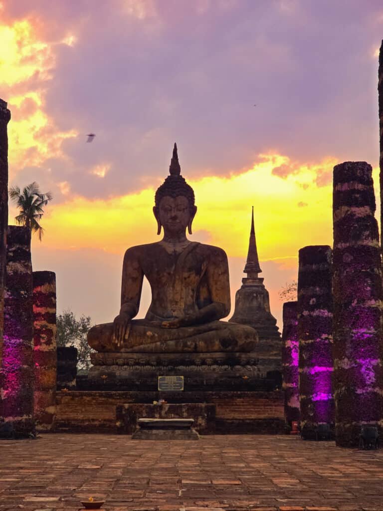 Statue de Bouddha au coucher du soleil dans un temple d’Asie, entourée de colonnes anciennes et de lumière dorée.