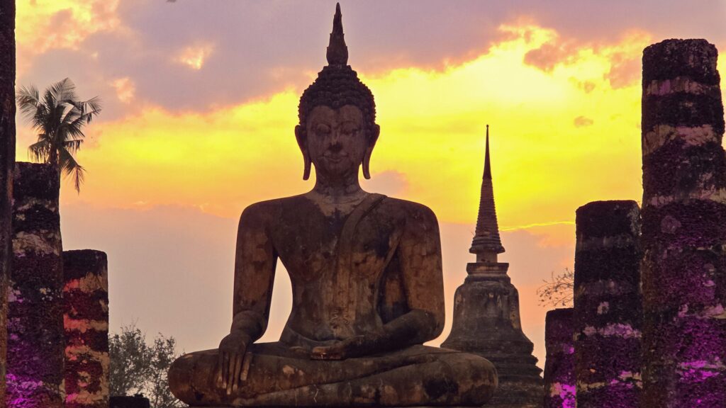 Statue de Bouddha au coucher du soleil dans un temple d’Asie, entourée de colonnes anciennes et de lumière dorée.