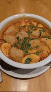 Bol de soupe Tom Yum thaïlandaise, bouillon orangé épicé au lait de coco, garni d’herbes fraîches et de boulettes.
