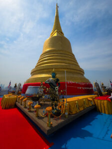 Stupa doré du Wat Saket à Bangkok, sous un ciel bleu, entouré de décorations et offrandes.