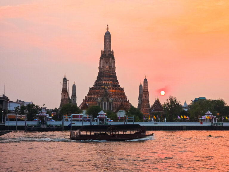 Le Wat Arun de Bangkok au coucher du soleil, vu depuis le fleuve Chao Phraya avec un bateau traditionnel au premier plan.