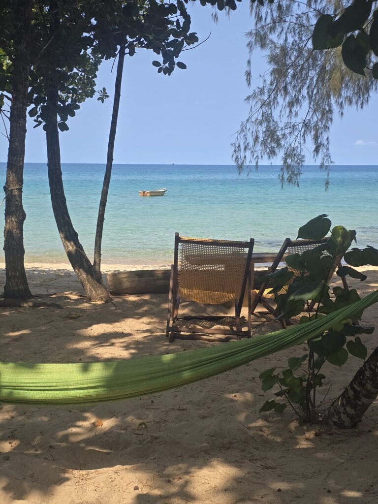 Hamac vert tendu entre deux arbres sur la plage de Sunset Beach à Koh Rong Samloem, avec deux transats en bois face à la mer turquoise et un petit bateau au large sous un ciel bleu clair.