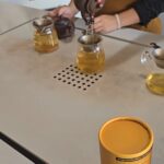 Gros plan sur une dégustation de thé à la plantation Choui Fong à Chiang Rai. Une main verse du thé d'une théière en terre cuite dans des tasses en verre transparent, avec un tube de thé Oolong jaune au premier plan.