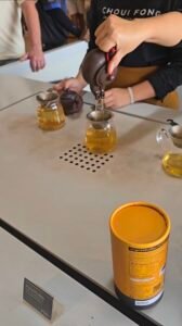 Gros plan sur une dégustation de thé à la plantation Choui Fong à Chiang Rai. Une main verse du thé d'une théière en terre cuite dans des tasses en verre transparent, avec un tube de thé Oolong jaune au premier plan.
