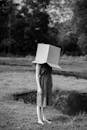 Black and white photo of a woman outdoors in nature, head hidden in a box.