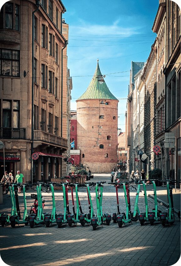 Trottinettes électriques vertes alignées en premier plan dans une rue pavée, avec la tour de la Poudre ronde en brique et son toit conique vert au fond, encadrée de façades anciennes